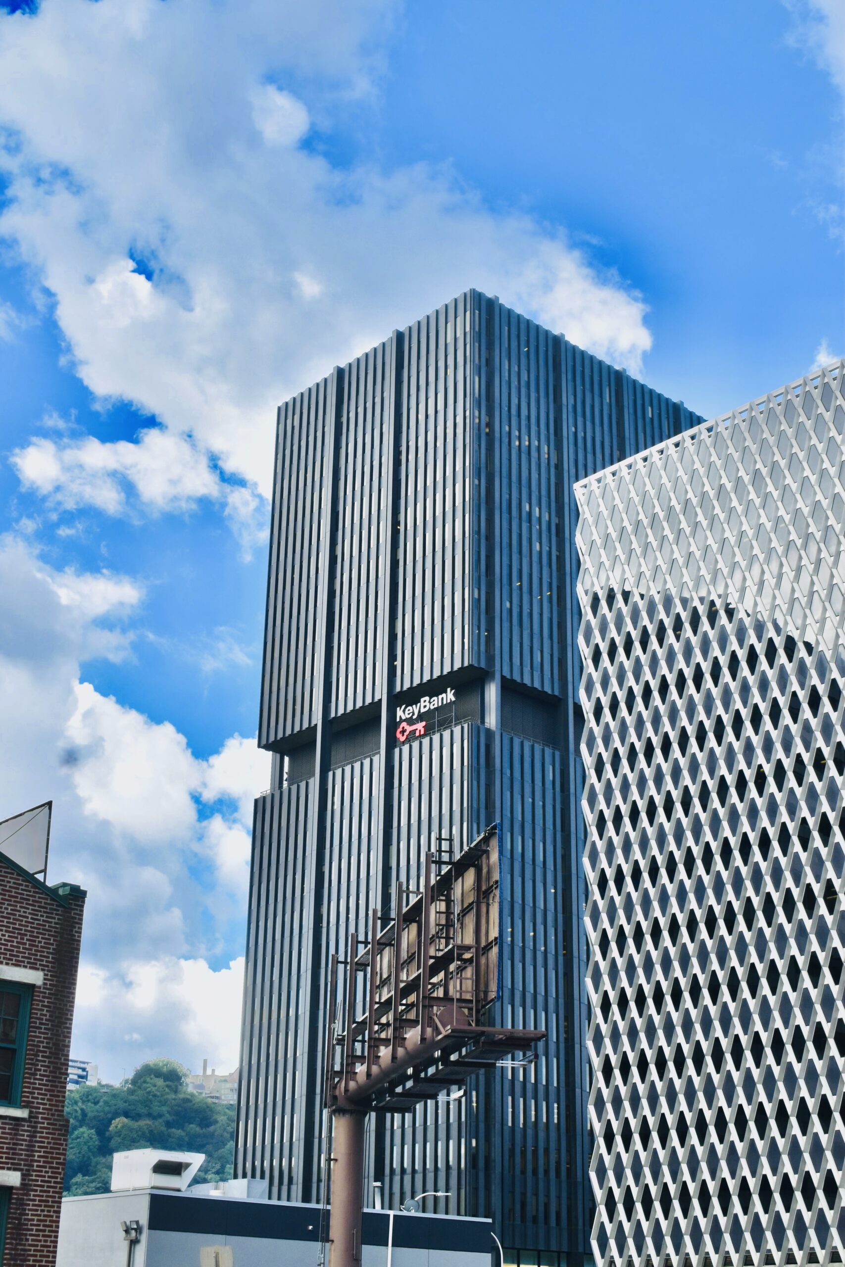 Two tall modern buildings against a blue sky with clouds. The left building features a large billboard. The right building has a unique textured facade, while the center building displays the KeyBank logo.