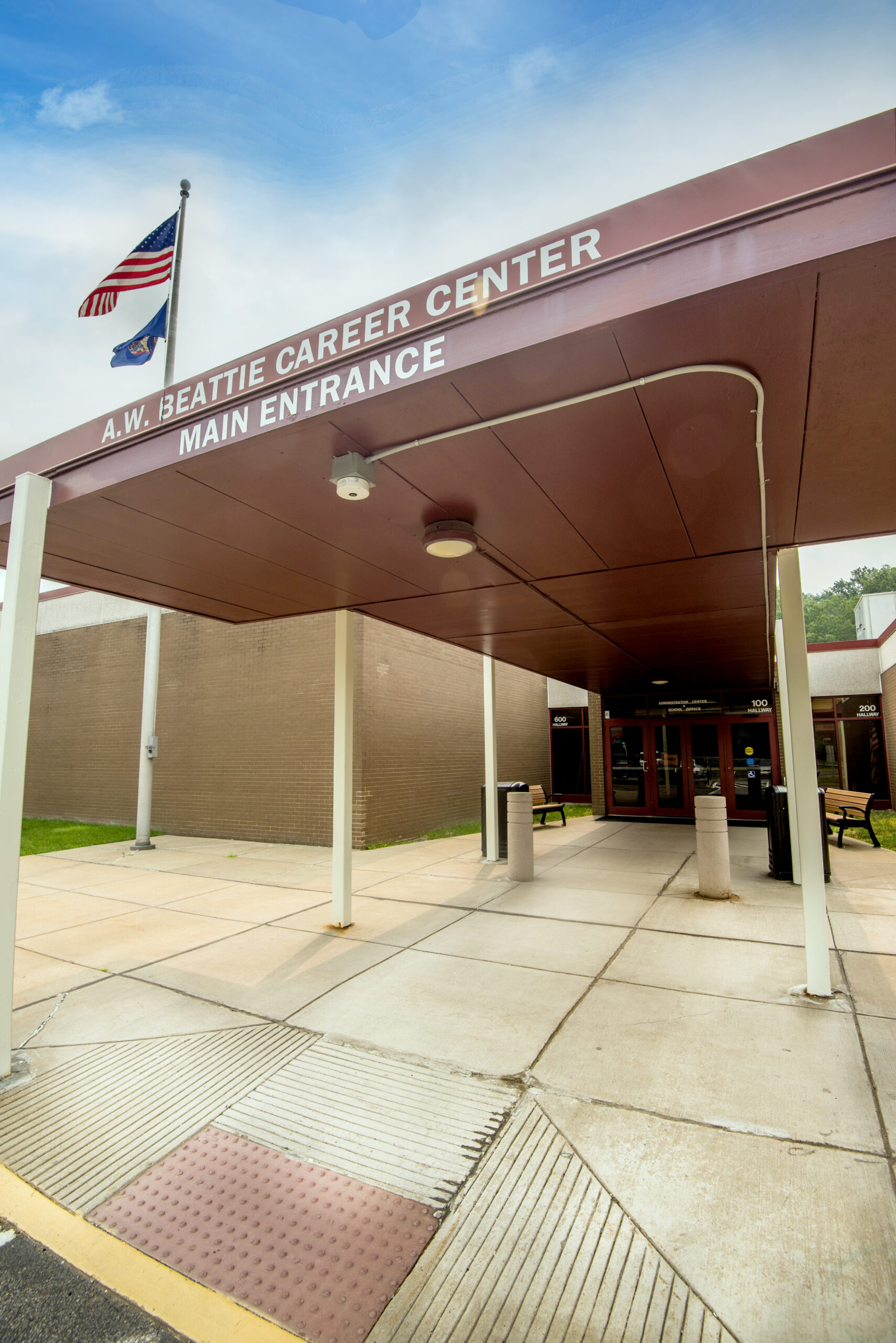 Exterior view of A.W. Beattie Career Center's main entrance, featuring a covered walkway and American flag. Sidewalk and benches are visible.