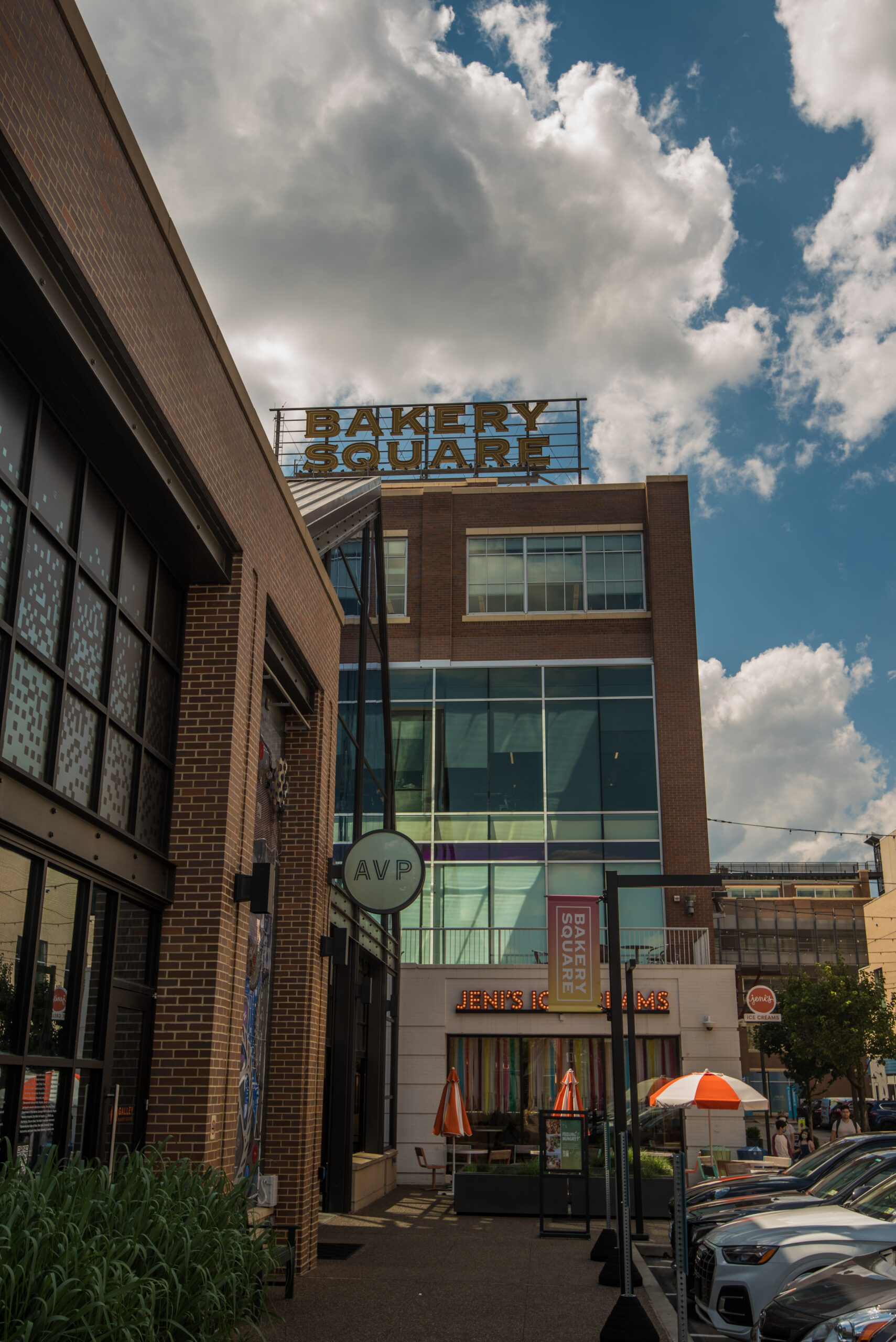 An outdoor area with a sign indicating "Bakery Square". Several parked cars line the street with modern buildings on either side. The sky is partly cloudy.