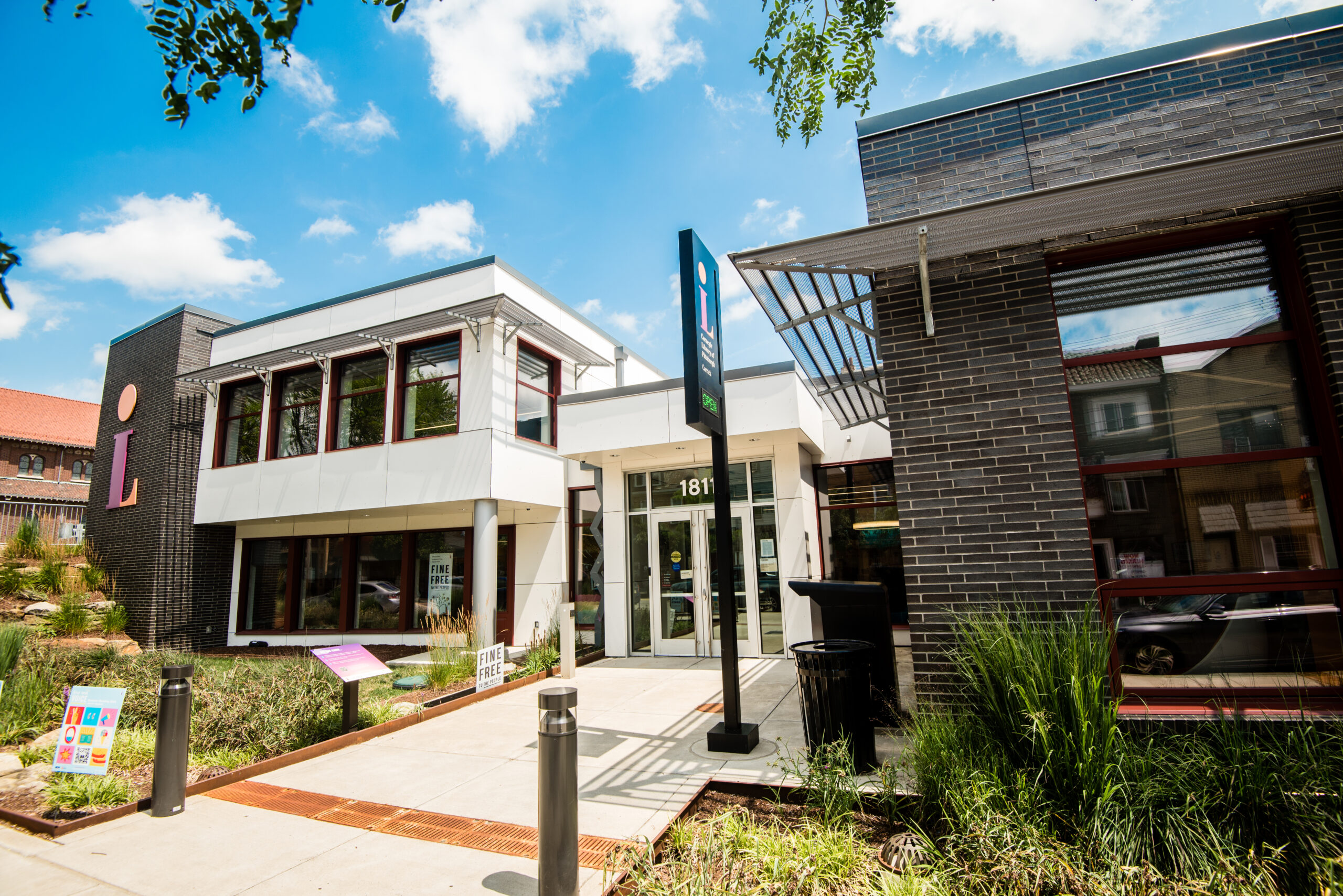 An exterior view of a modern building with large windows and a flat roof. The entrance features glass doors, with a sign displaying the number 181. Surrounding the building are green plants and landscaping under a clear blue sky.