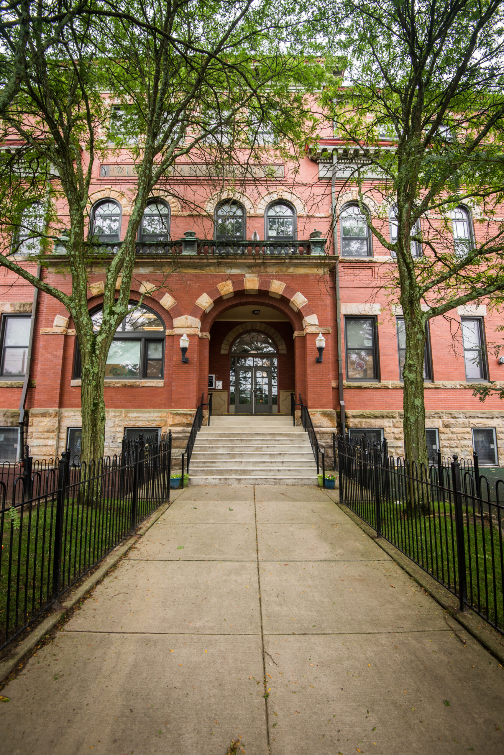A brick building with large steps leading to the entrance. Two trees flank the pathway. The facade has large windows and decorative details. A black fence surrounds the grassy area.