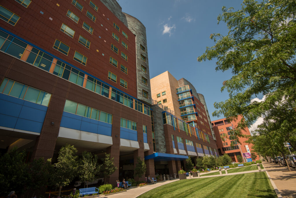 Childrens Hospital of Pittsburgh exterior. A view of a modern multi-story building featuring a mix of red and beige facades. Lush green grass and trees are in the foreground, with people seated outside on benches. The sky is clear with a few clouds.