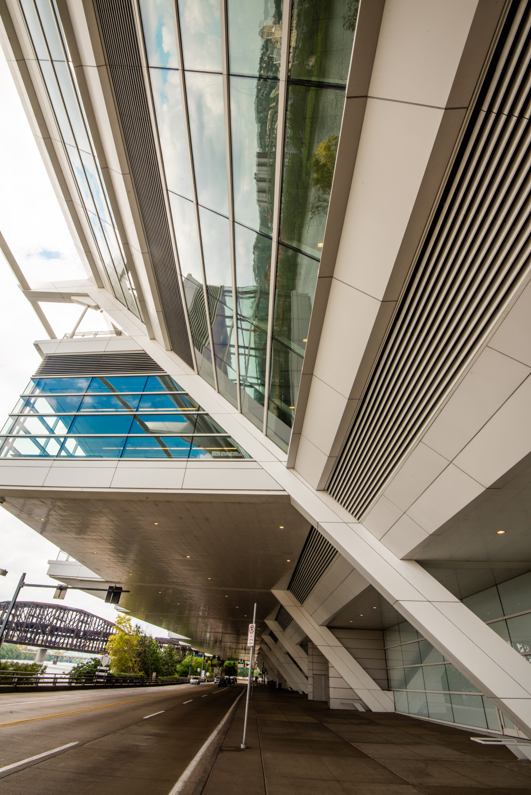 A modern building with a glass facade and angular architecture, viewed from a low angle. The structure reflects elements of the sky and nearby greenery. A roadway is visible in the foreground.