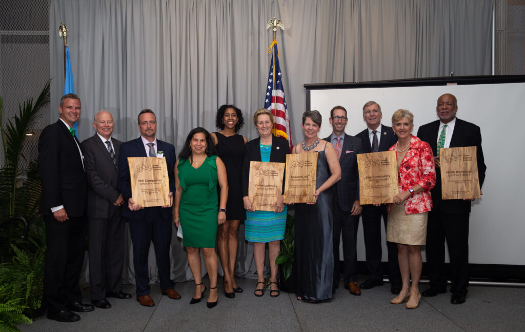 A group of professionals standing in front of a screen, holding wooden plaques. They are in a formal setting with a U.S. flag and green plants in the background. The group includes men and women in business attire.