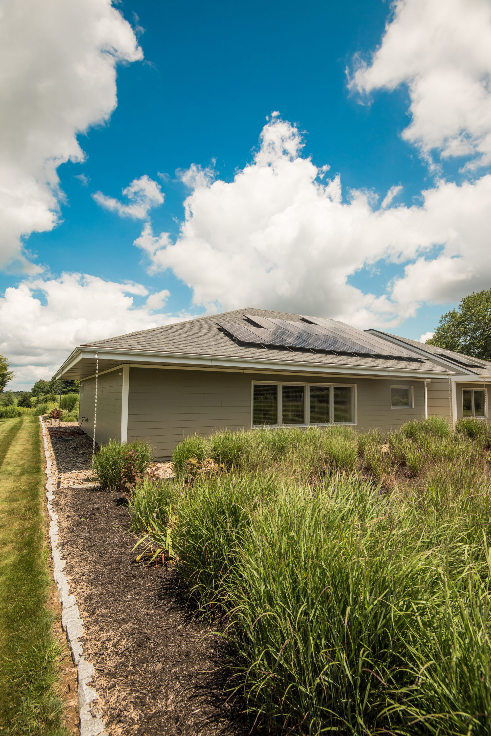 A modern building with solar panels on the roof, surrounded by grass and shrubs, under a partly cloudy sky. The building features a light-colored exterior and large windows.