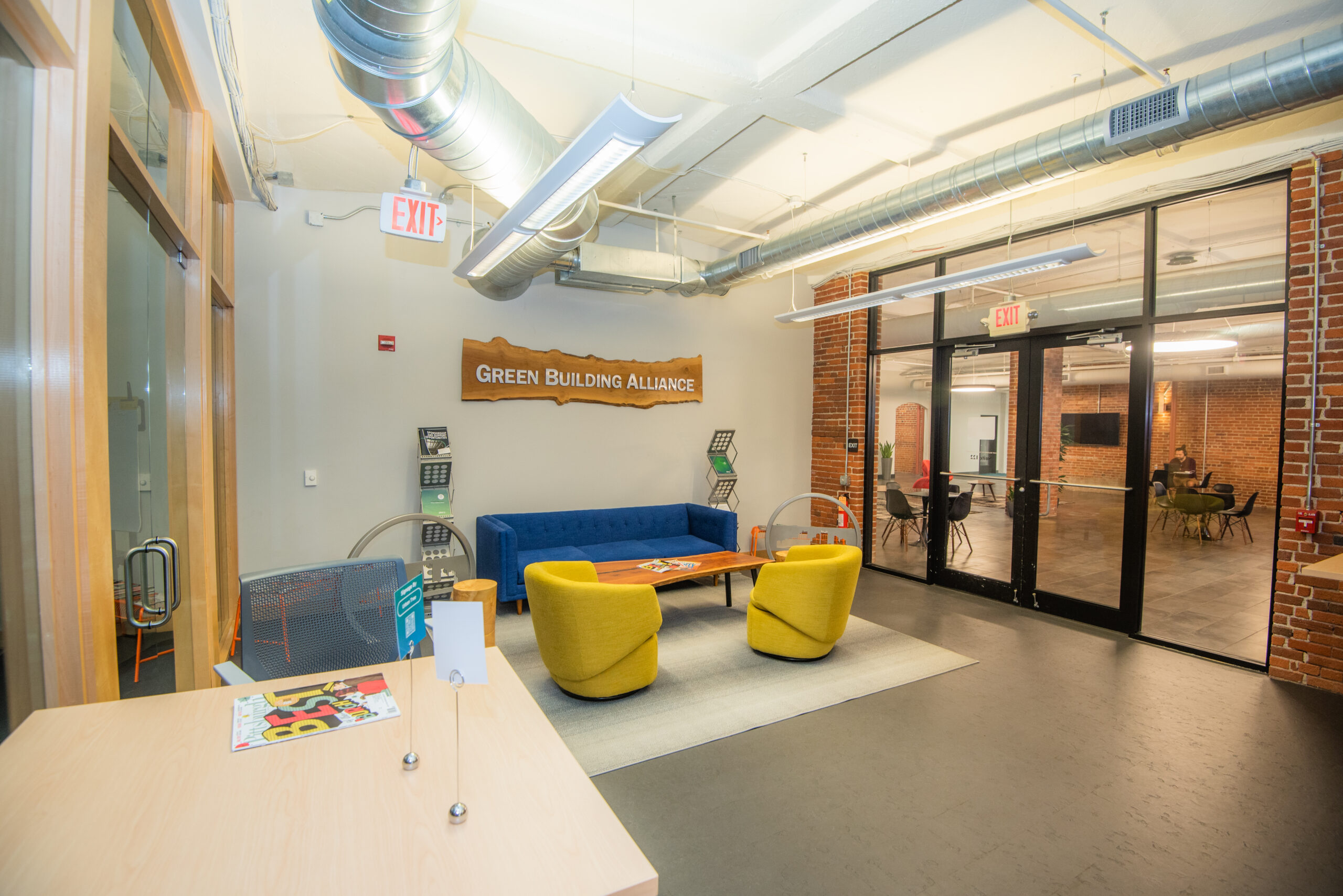 A modern office lobby featuring a blue sofa and two yellow chairs around a wooden coffee table. A sign reads "Green Building Alliance" on the wall. There are glass doors leading to another room and a reception desk on the left.