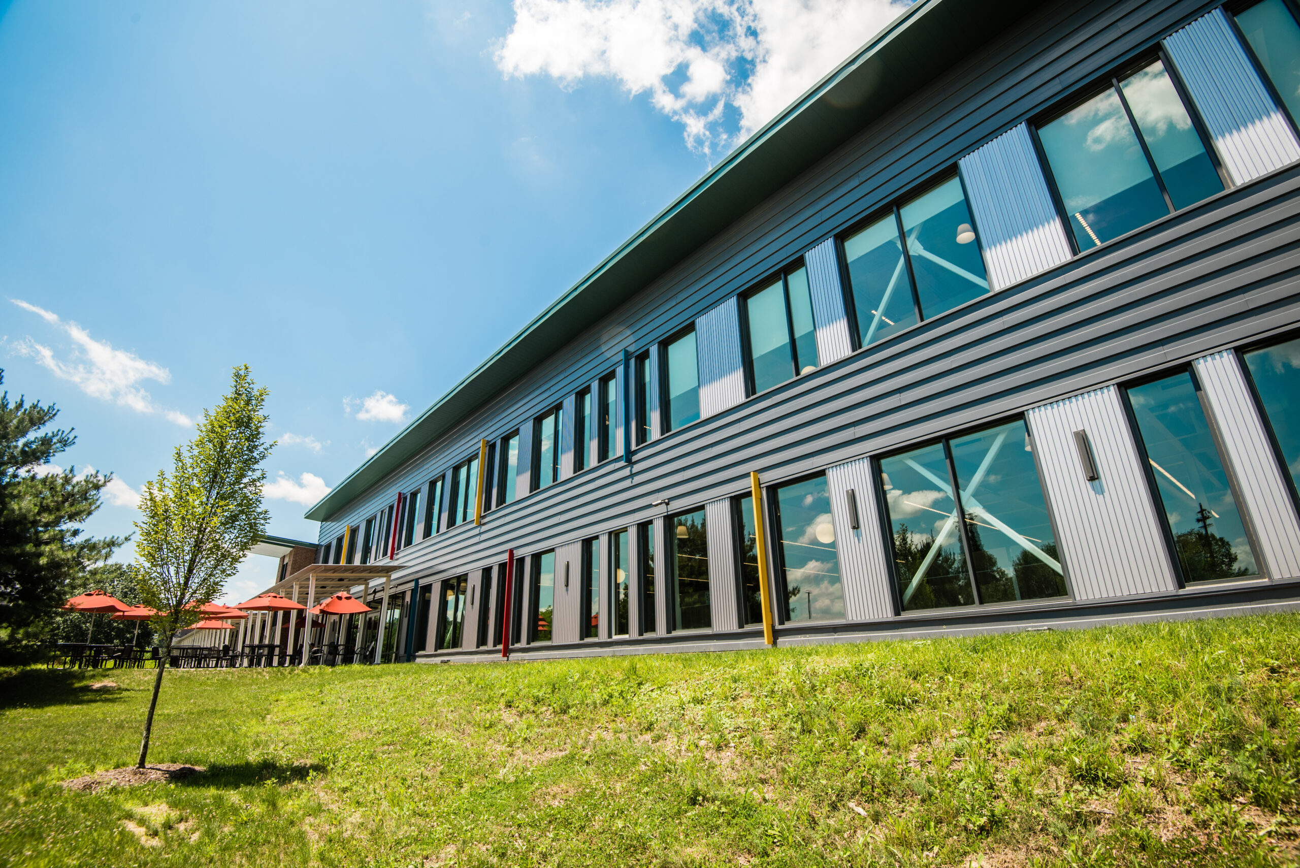 A modern building with large windows and grey siding, viewed from an angle. The building features red accents and a green lawn in front. A small tree is visible, along with outdoor seating under red umbrellas.