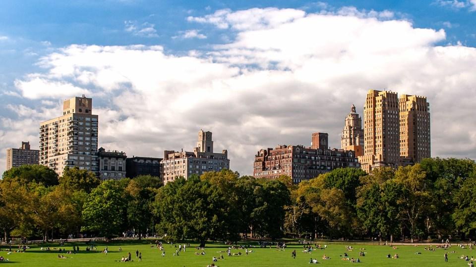 A panoramic view of a park with green grass and scattered people, surrounded by various buildings in the background under a partly cloudy sky.