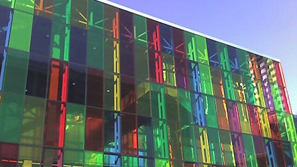 A modern building facade with colorful glass panels in green, red, and yellow reflecting light. The structure features a geometric design with visible support beams and a clear sky in the background.