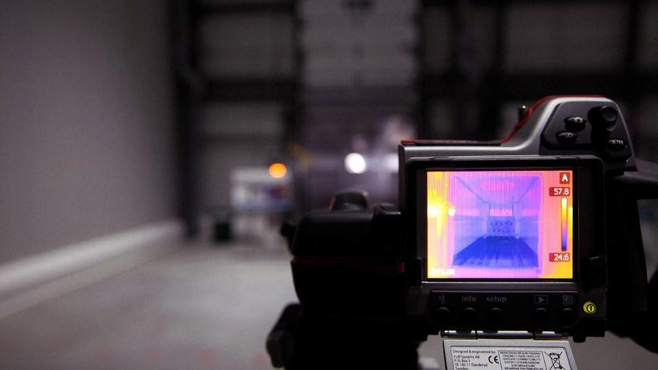 An infrared camera focused on an industrial space, displaying a thermal image on its screen. The image shows varying temperatures, with prominent colors indicating heat levels. A gray wall and equipment are visible in the background.