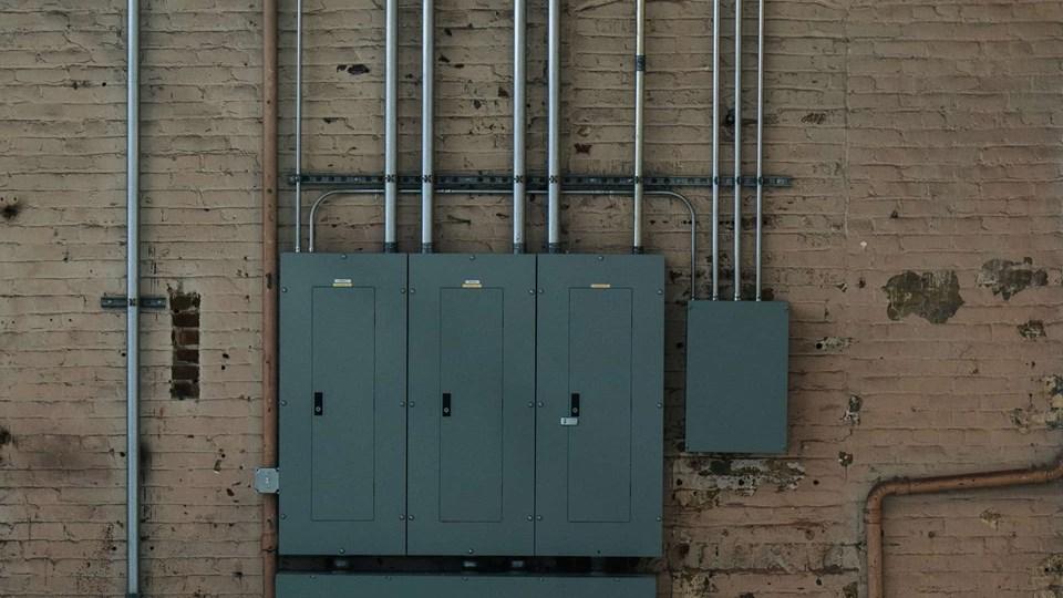 Three gray electrical panels mounted on a brick wall, with metal conduits and wiring extending from the panels. The wall shows signs of wear and discoloration.