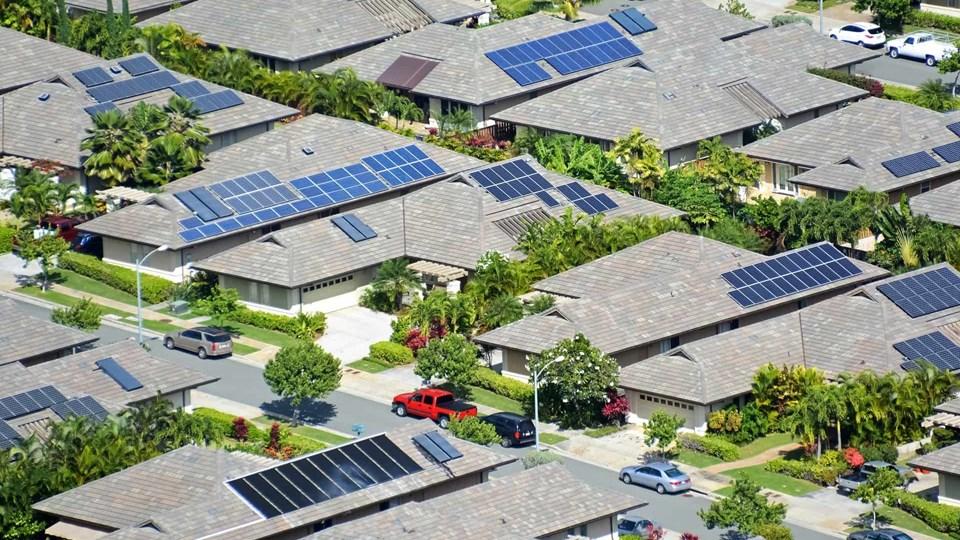 An aerial view of houses with solar panels on their roofs in a residential neighborhood. Lush greenery and parked cars are visible along the streets.