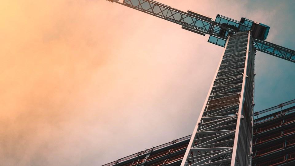 A tall construction crane extending upward against a colorful sky. The crane is near a building under construction, with metal beams visible.