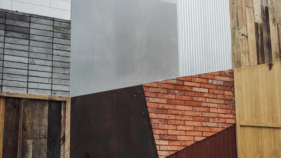 A textured wall composed of various materials, including wooden planks, metal sheets, and brick. The arrangement features sections of gray wood, bright red bricks, and metallic surfaces creating geometric shapes.