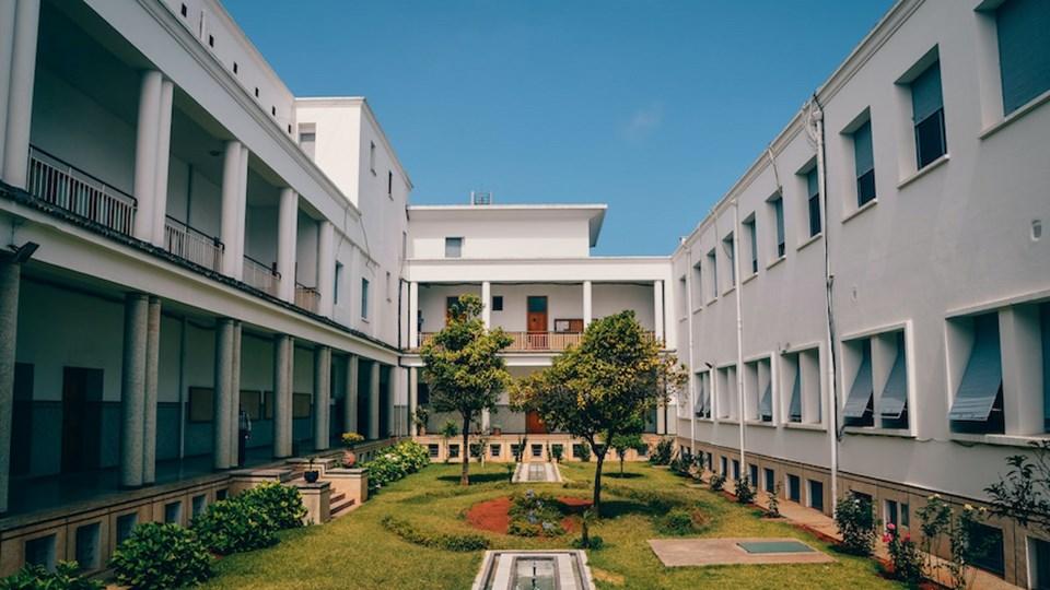 A courtyard surrounded by white buildings with multiple windows. Grass and small trees are visible in the center, along with a stone path leading to a fountain area. The sky is clear and blue, indicating a sunny day.