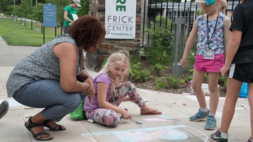 A woman crouches beside a girl drawing with chalk on a sidewalk. Three children are engaged in creative activities outside the Frick Environmental Center, with green spaces visible in the background.