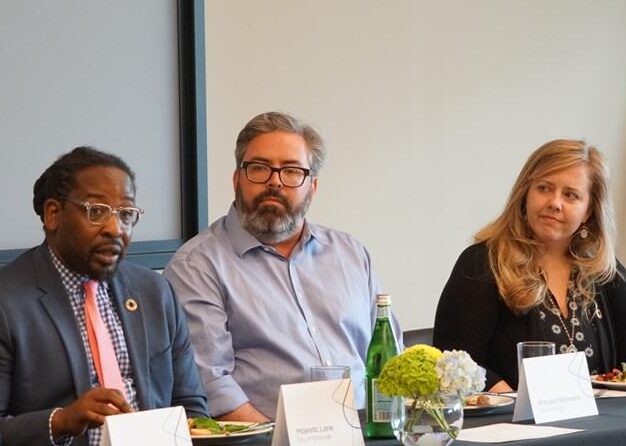 Three professionals seated at a table during a discussion. The man on the left is wearing glasses and a patterned tie, the man in the center has a beard and wears a light blue shirt. A woman on the right has long hair and is wearing a black jacket.