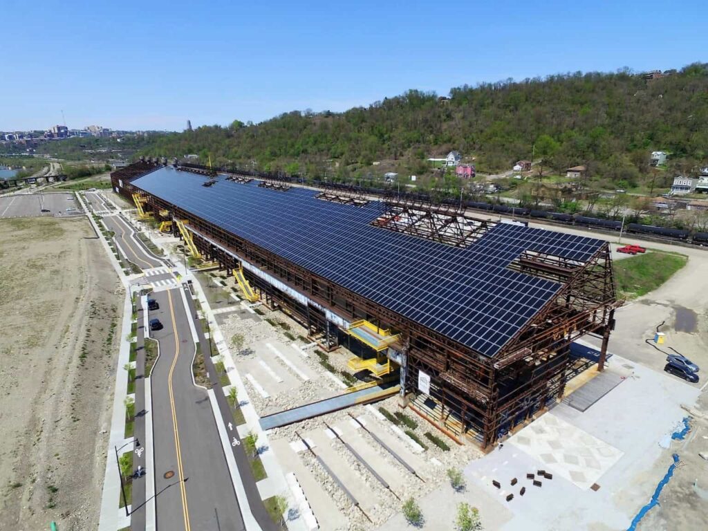 A large commercial building under construction with a solar panel roof. Surrounding area features bare land and paved roads, with trees on a hillside in the background.