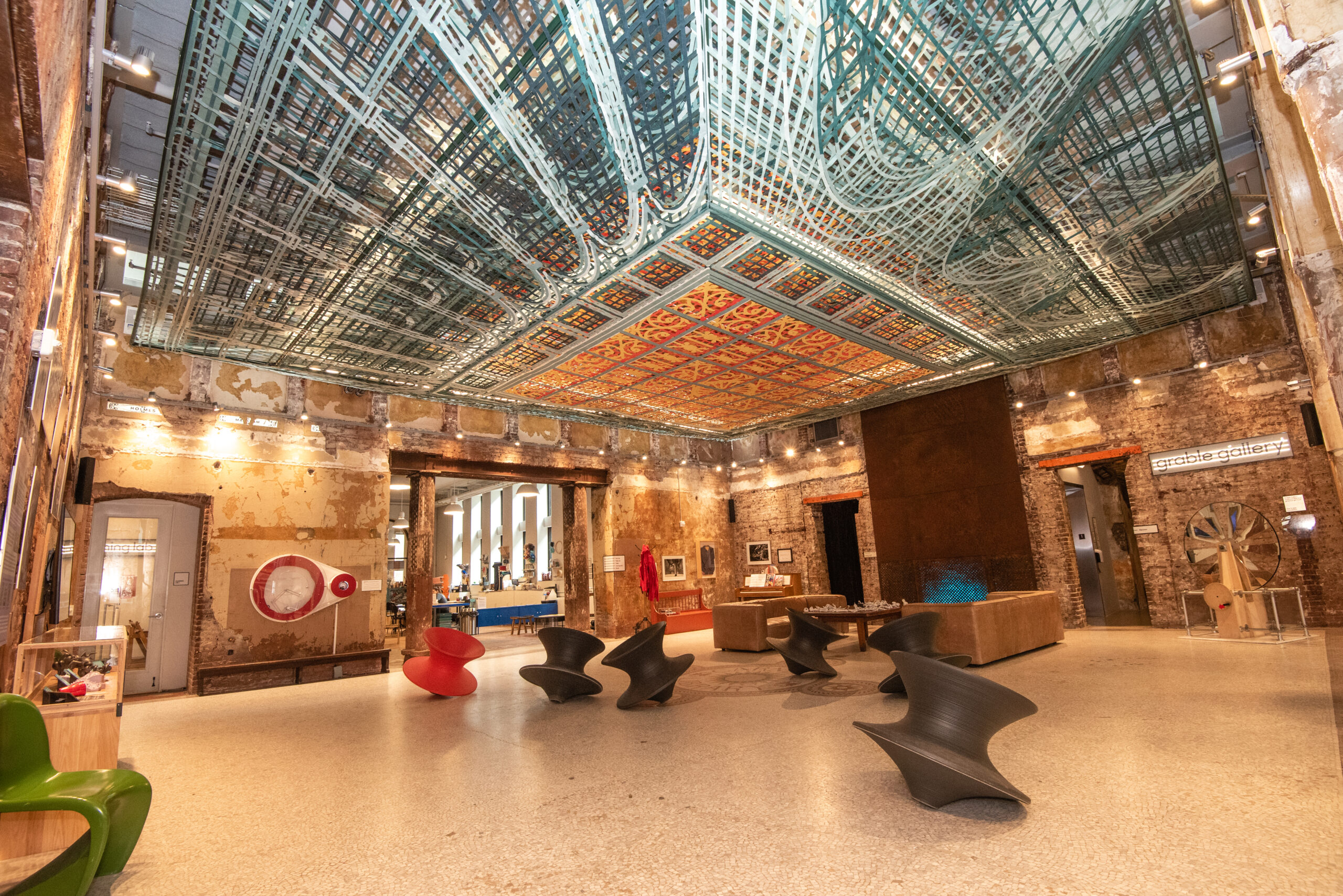 MuseumLab lobby. A spacious lobby featuring a colorful, ornate ceiling with intricate designs. Stylish black and red chairs are arranged on a light-colored floor. Exposed brick walls are seen, along with modern decor and art on display.