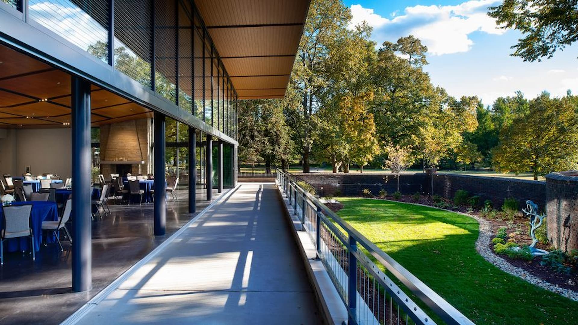 Exterior view of a modern building with large glass windows, showcasing a walkway leading to a green lawn and trees. Tables are set up inside the building, with blue tablecloths visible.