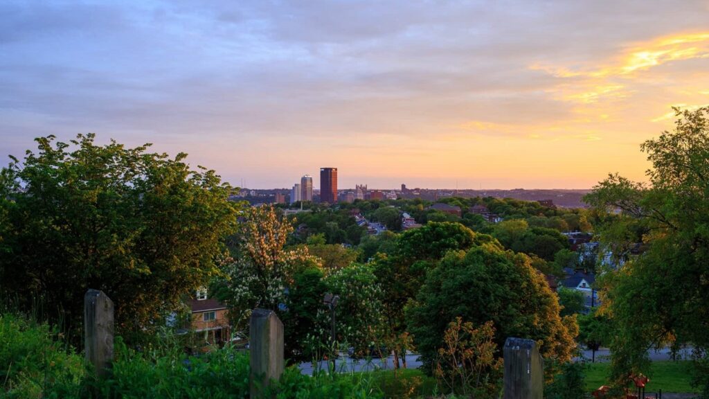 A view of the Pittsburgh skyline at sunset, with buildings in the background and greenery in the foreground. The sky displays shades of orange and blue, highlighting the horizon.