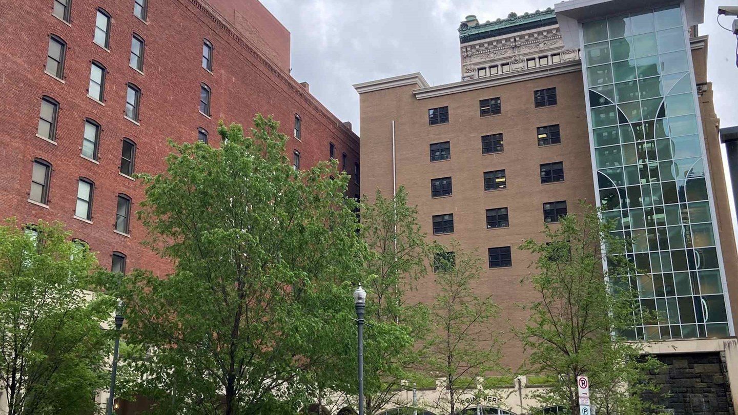 View of several buildings with brick facades and glass elements, surrounded by trees. The scene features a mixture of architectural styles, highlighting urban development.