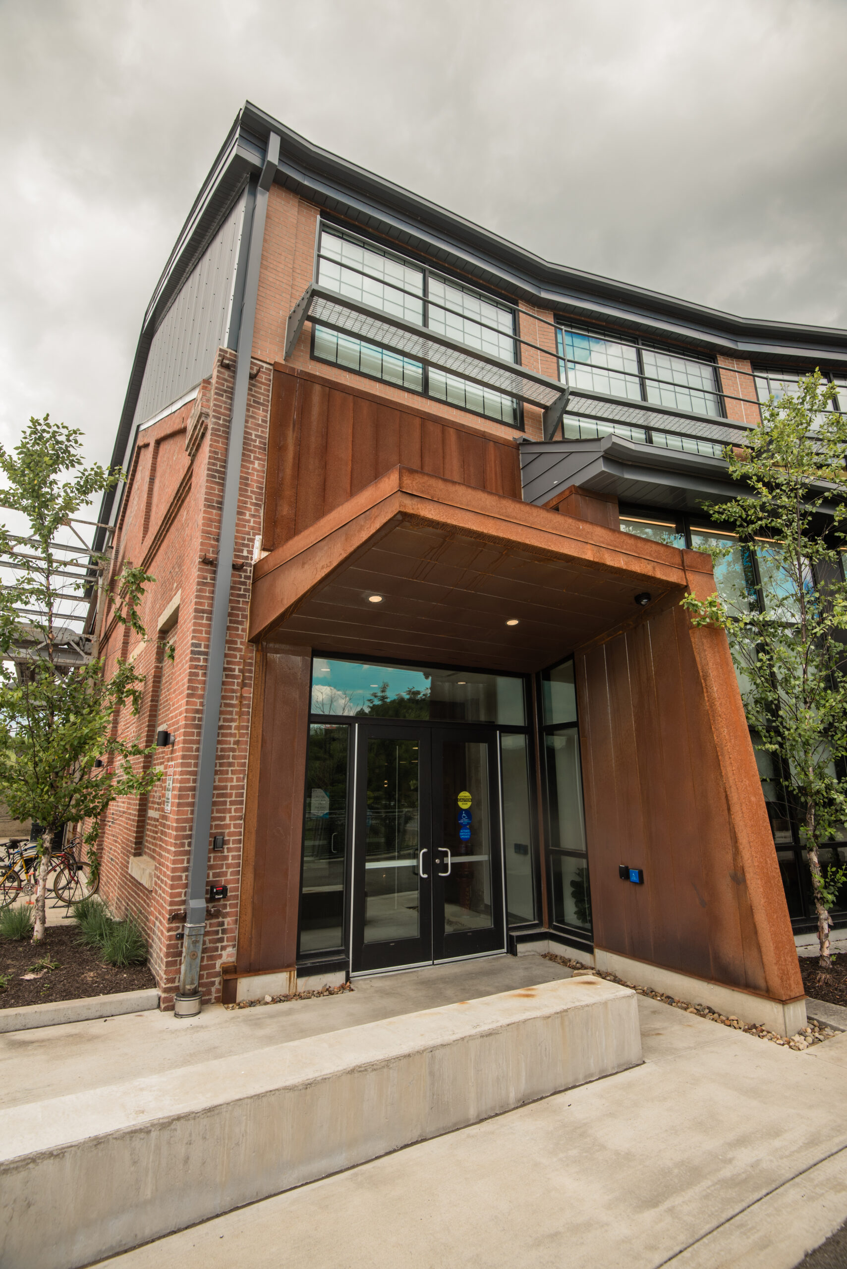 The entrance of a modern building featuring a copper-colored overhang, large glass windows, and landscaped greenery on either side of the entrance.