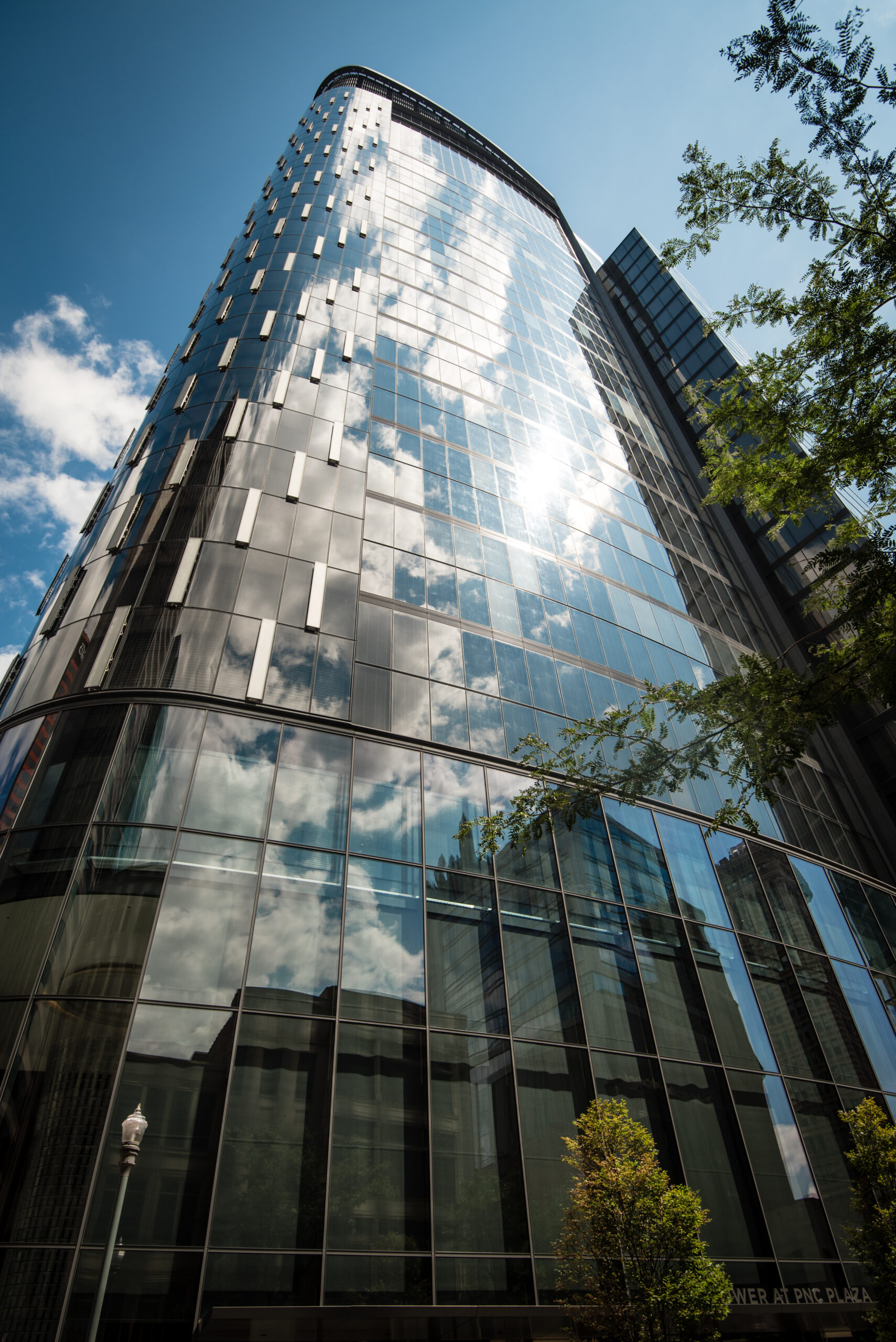 A modern glass building with a curved facade reflects clouds and blue sky. Trees are visible in the foreground, providing green accents to the architectural scene.