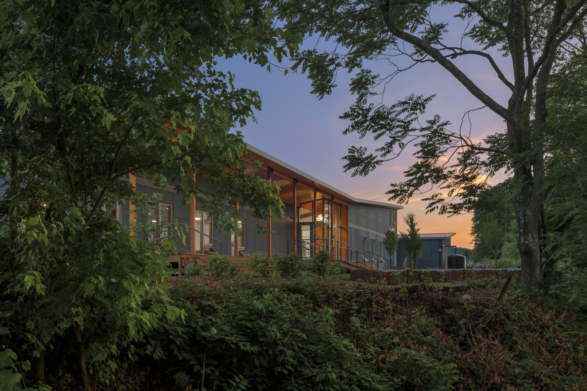 A modern building with wooden beams and large windows, surrounded by trees and greenery at dusk. The sky shows hints of sunset colors. A deck is visible in front of the building.