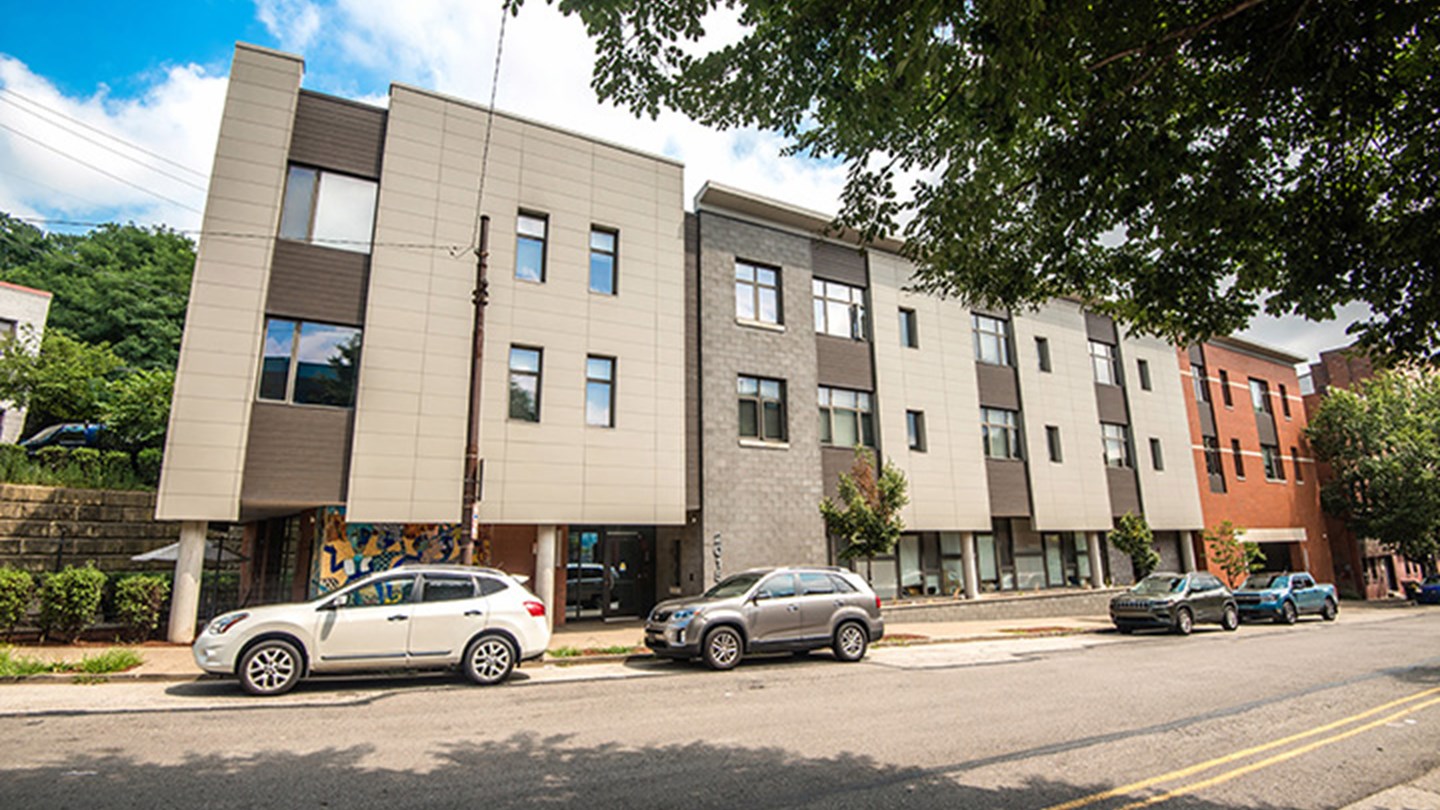 Modern multi-story residential building with a mix of light and dark exterior materials. Several parked cars are along the street and trees are visible in front of the building.