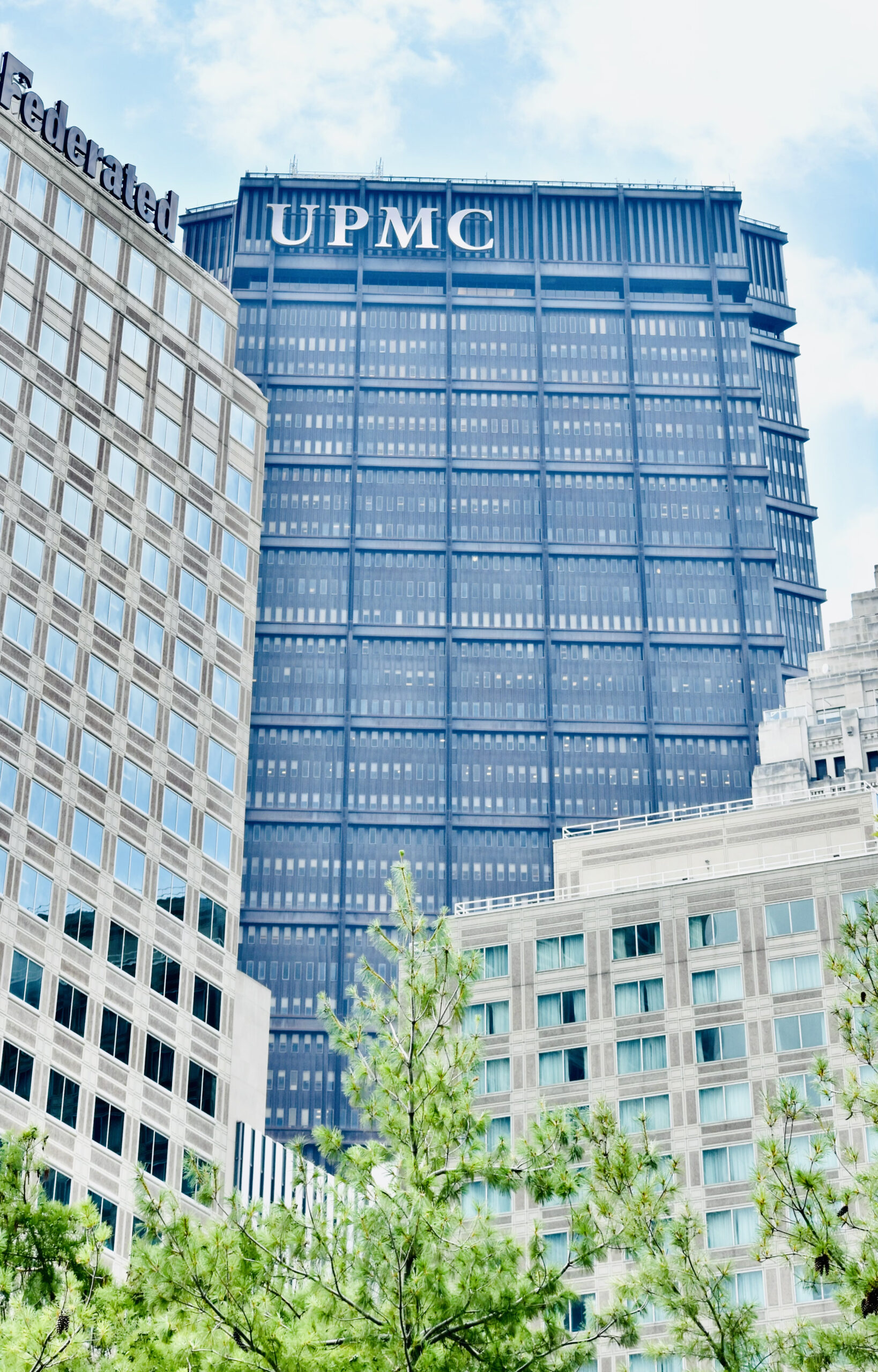 A tall blue skyscraper with the name 'UPMC' displayed at the top, surrounded by other buildings and green trees in the foreground. The sky is clear with some clouds.