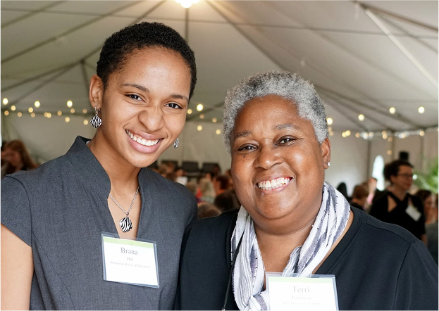 Two women smiling for a photo at an event under a tent. The woman on the left is wearing a dark gray outfit and has short curly hair. The woman on the right is wearing a black top with a white scarf and has gray hair.